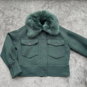 Mörkgrön jacka med fuskpäls - Säljer en superfin mörkgrön jacka från  meotine . Den har en lyxig fuskpäls på kragen som ger en extra touch av stil. Jackan är kort och har knappar framtill samt två fickor med lock. Perfekt för höst och vinter!