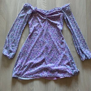 Lila blommig offshoulder klänning från NLY One - Säljer en superfin lila offshoulder klänning från NLY One i storlek XS. Klänningen har ett blommigt mönster i olika nyanser av lila och blå. Den är långärmad och har söta volangdetaljer. Materialet är mestadels polyester med lite elastan för en bekväm passform. Perfekt för vår och sommarens fester! Använd endast 1 gång.