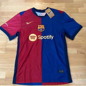 Säljer en FC Barcelona fotbollströja i röd och blå med korta ärmar. Tröjan har klubbens emblem och Spotify-logga på framsidan. På baksidan står det 'Lamine Yamal' och nummer 27. Tröjan är helt ny med etikett kvar.