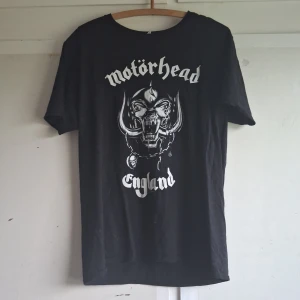 Svart Motörhead t-shirt från Gildan - Säljer en svart Motörhead t-shirt från Gildan. Den är i storlek L och är gjord av 100% bomull. På framsidan finns ett coolt tryck med Motörhead-loggan och en bild av deras ikoniska maskot. På baksidan står det 'Everything Louder Than Everything Else' i vit text. Perfekt för alla rockfans! 🤘
