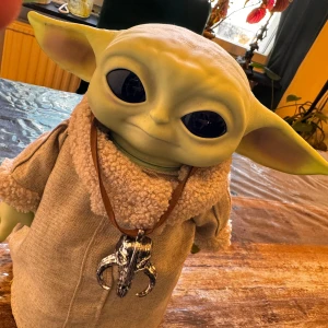 Baby Yoda docka med beige kläder - Säljer en supergullig Baby Yoda docka med stora svarta ögon och gröna öron. Dockan har på sig en beige klädsel med fluffiga detaljer runt kragen och ärmsluten. Den har också ett halsband med en silverfärgad berlock. Perfekt för alla Star Wars-fans!