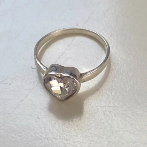 Silverring med hjärtformad sten - Säljer en vacker silverring med en stor, glittrande hjärtformad sten. Ringen är i bra skick och har en elegant och tidlös design. Perfekt för speciella tillfällen eller som en fin present. Märkt med 'Sasb' och är inte magnetisk så jag tror det är silver 