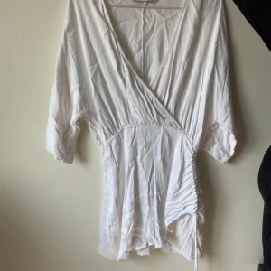 Vit omlottklänning från Zara - Säljer en superfin vit omlottklänning från Zara. Den har trekvartsärmar och en snygg rynkad detalj på sidan. Perfekt för sommaren eller en festlig kväll! Klänningen är i ett lätt och bekvämt material.