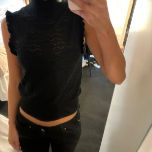 Svart ärmlös topp med volangdetaljer - Säljer en snygg svart ärmlös topp med hög krage och volangdetaljer på axlarna. Toppen har ett fint mönster och är perfekt för både vardag och fest.