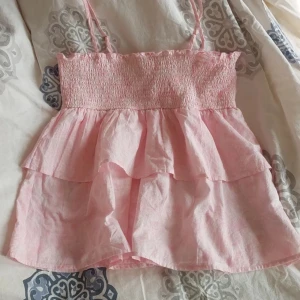 Rosa volangtopp från Gina Tricot - Säljer en supersöt rosa topp från Gina Tricot med mönster. Den har smala axelband som man kan ta av och på och en smockad överdel som ger en skön passform. Toppen är dekorerad med volanger. Den är aldrig använd och Perfekt för sommardagar eller en fe