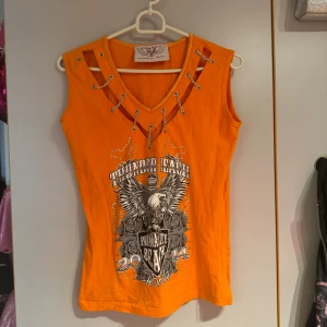 Orange linne/topp  - Cool topp med tryck och strass! Köpt second hand men aldrig använd. Saknar en kedja. Står ej storlek men skulle säga M (jag är L/XL och den sitter tight på mig) 
