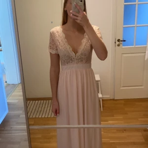 Ljusrosa långklänning med spetsdetaljer - Säljer en superfin ljusrosa långklänning med vackra spetsdetaljer på överdelen. Klänningen har korta ärmar och en djup v-ringning både fram och bak som ger en elegant look. Den är perfekt för fester eller bal. Klänningen har en dragkedja i ryggen. använd en gång så ser ut som ny! 