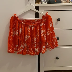 Orange blommig offshoulder topp från BikBok - Säljer en superfin orange offshoulder topp från BikBok med vita blommor. Den har en elastisk kant upptill och vid midjan för en bekväm passform. Perfekt för sommardagar eller en festlig kväll!
