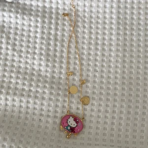 Guldigt halsband med Hello Kitty-berlock - Säljer ett supergulligt guldigt halsband med en stor Hello Kitty-berlock. Berlocken är rosa med en söt Hello Kitty-figur i mitten och små hängande guldklockor. Kedjan har också små stjärnor och runda plattor som detaljer. Perfekt för alla Hello Kitty-fans! 🐱💖