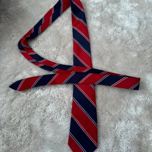 Randig slips i rött och blått - en randig slips i rött och blått. Den är gjord av 100% polyester och tillverkad i Hong Kong. 