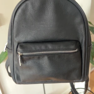 Svart miniryggsäck med dragkedja - Säljer en stilren svart miniryggsäck i skinnimitation. Den har en praktisk dragkedja framtill och justerbara axelremmar. Perfekt för vardagsbruk eller en dag på stan. Ryggsäcken är i bra skick och redo för nya äventyr!