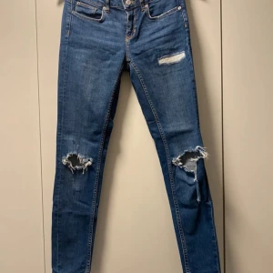 Blå slitna jeans - Säljer ett par blå jeans med snygga slitningar på knäna och låren. De har en tight passform och är perfekta för en avslappnad stil. Jeansen har en klassisk femficksdesign och är i ett välanvänt skick. Perfekta för både vardag och fest!