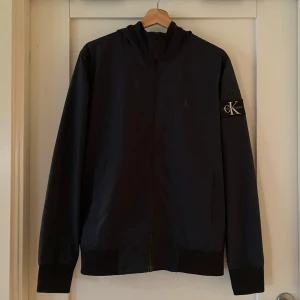 Calvin Klein Jeans mörkblå jacka, storlek S - Helt oanvänd jacka med huva från Calvin Klein Jeans. Jackan har en dragkedja framtill och två fickor på sidorna. Det finns en liten logga på bröstet och en större logga på vänster ärm.  Inga skador eller inget bekymmer på jackan,  ny pris 1800 kr