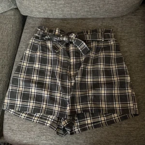 Rutig shorts med bälte - Snygga högmidjade shorts i svart och beige rutigt mönster. De har ett matchande bälte som ger en extra touch. Perfekta för en stilren look. Det står ingen storlek i dem, men uppskattningsvis är de storlek M.