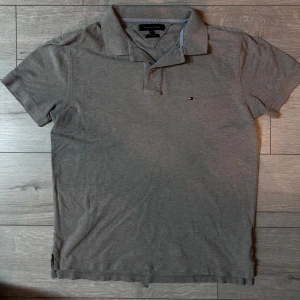pikétröja från Tommy Hilfiger - Säljer en stilren grå pikétröja från Tommy Hilfiger. Tröjan har en klassisk krage och korta ärmar, med den ikoniska logotypen broderad på bröstet. Perfekt för en avslappnad men ändå snygg look.