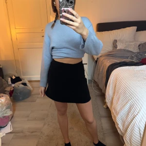 Svart kortkjol med knappar - Snygg svart kortkjol med knappar framtill. Perfekt för en stilren look. Kjolen har en enkel design som gör den lätt att matcha med olika toppar. Mocka tyg, ser fläckig ut i ljuset men det är den inte!!!! Den är köpt för länge sen!! 