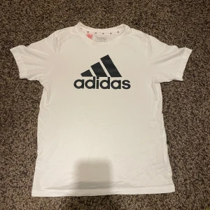 Vit t-shirt från Adidas - Säljer en klassisk vit t-shirt från Adidas med deras ikoniska logga i svart på bröstet. Perfekt för en sportig look eller avslappnad stil. Kortärmad och bekväm passform.