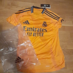 Orange fotbollströja från Adidas - Säljer en orange fotbollströja från Adidas med korta ärmar och svarta detaljer. Tröjan har Real Madrids emblem och sponsorlogga på framsidan samt spelarnamnet 'Mbappé' och nummer 9 på baksidan. Perfekt för fotbollsfans! 🧡 årsmodell 2025 borta