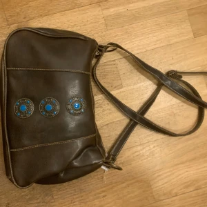 vintage shouderbag - Snygg brun axelväska i skinn med tre dekorativa blå detaljer på framsidan. Väskan har en praktisk dragkedja på baksidan och en justerbar axelrem för bekväm passform.