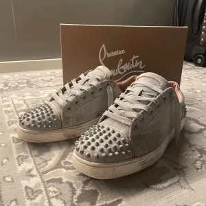 Grå sneakers med nitar från Christian Louboutin - Snygga grå sneakers från Christian Louboutin med ikoniska röda sulor. Skorna har ett unikt utseende med nitar över hela ovandelen och snörning framtill. Perfekta för att ge din stil en edgy touch. Pris kan diskuteras!
