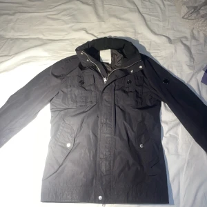 Svart jacka från J.Lindeberg - Snygg svart jacka från J.Lindeberg med dragkedja och knappar. Jackan har en stilren design med praktiska fickor och en hög krage. Perfekt för en stilren look. Jackan är i strolek S och nypris ligger på ungefär 4000-3500. Jackan går inte få tag på längre och har ingen användning utav den då jag väljer att sälja den.