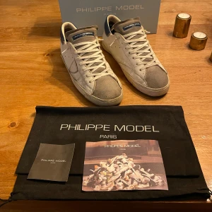 Sneakers från Philippe Model - Snygga sneakers från Philippe Model i en kombination av vitt och grått med blå detaljer. Skorna har snörning och en stilren design med märkets namn på sulan. Perfekta för en avslappnad stil. Tvättade skorna innan så mockan ser lite blöt ut. Precis är fast 1600 är lägsta jag går. Om du har några funderingar så tveka inte på att fråga mig🔥