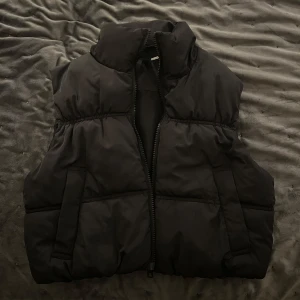 Svart dunväst från H&M Divided - Säljer en svart dunväst från H&M Divided. Västen har en dragkedja framtill och en hög krage för extra värme. Perfekt för kyliga dagar när du vill hålla stilen enkel och bekväm.