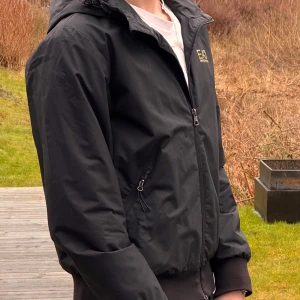 Svart jacka från EA7 Emporio Armani - Snygg svart jacka från EA7 Emporio Armani med dragkedja och huva. Jackan har långa ärmar och ribbade muddar. Perfekt för en stilren look.