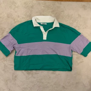 Grön och lila pikétröja från Pull&Bear - Säljer en cool grön och lila pikétröja från Pull&Bear. Tröjan har en klassisk krage och korta ärmar. Perfekt för en avslappnad stil.
