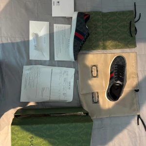 Gucci sneakers med mönster - Snygga Gucci sneakers i grått med ett distinkt mönster. Skorna har klassiska röda och blå ränder på sidan och svart snörning. Perfekta för en stilren look.