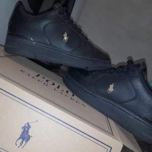 Polo Ralph Lauren Skor - Snygga svarta sneakers från Polo Ralph Lauren med guldlogga på sidan och hälen. Skorna har en klassisk design med snörning och är tillverkade i skinn. Perfekta för en stilren look. TVEKA INTE PÅ ATT FRÅGA! Kan skicka mer bilder. 