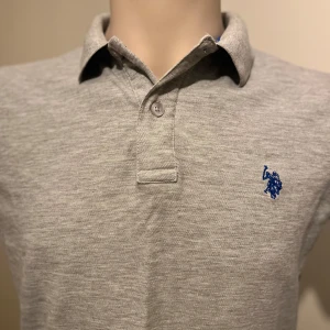 Grå pikétröja från U.S. Polo Assn. - Säljer en stilren grå pikétröja från U.S. Polo Assn. med klassisk krage och knappslå. Tröjan har en broderad blå logotyp på bröstet och är tillverkad i premium bomull för extra komfort. Perfekt för en avslappnad look.