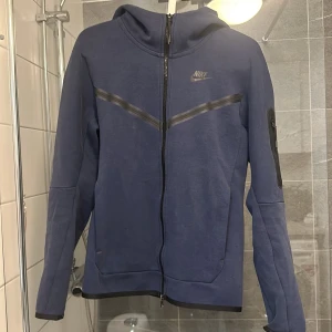 Blå hoodie från Nike - Säljer en blå hoodie från Nike med dragkedja och huva. Jackan har långa ärmar och en diskret Nike-logga på bröstet. Perfekt för en sportig look eller avslappnade dagar. Pris kan diskuteras 