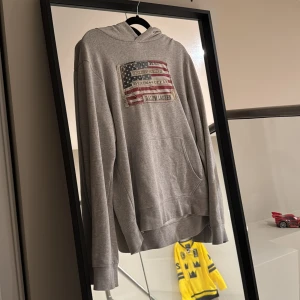 Grå hoodie från Denim & Supply Ralph Lauren - Säljer en grå hoodie från Denim & Supply Ralph Lauren med ett coolt amerikanskt flaggmönster på bröstet. Tröjan har en klassisk känguruficka och långa ärmar. Perfekt för en avslappnad stil. Vid frågor eller funderingar är det bara att höra av sig💯