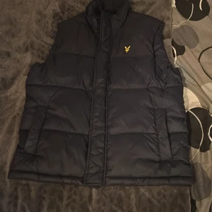 Svart dunväst från Lyle & Scott - Säljer en svart dunväst från Lyle & Scott med en gul broderad logotyp på bröstet. Västen har en hög krage och stängs med dragkedja. Perfekt för kyliga dagar