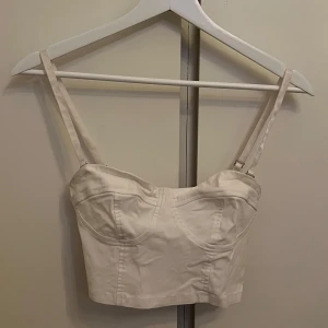 Vit topp - Snygg vit korsettopp med justerbara axelband (går även att ta av banden). Perfekt för att skapa en stilren look. Toppen har en figurnära passform och är tillverkad i ett mjukt material.