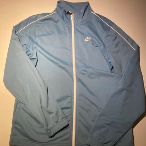Ljusblå tröja från Nike - Snygg ljusblå tröja från Nike med dragkedja och vita detaljer. Perfekt för träning eller som en lätt tröja till vardags. Den har långa ärmar och en hög krage för extra skydd. Mycket bra skick. Passar både L och M