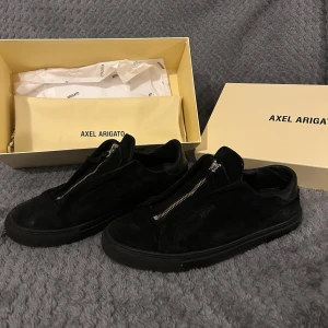 Svarta sneakers från Axel Arigato - Snygga svarta sneakers från Axel Arigato med dragkedja på ovansidan. Skorna är tillverkade i mocka och har en stilren design.  Har ett litet hål i hälen (sista bilden) men inget man märker av riktigt. Annars väldigt bra skick 