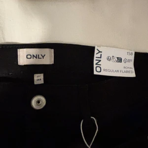 Svarta byxor från ONLY - Snygga svarta byxor från ONLY i modellen Royal Regular Flared. Perfekta för en stilren look med en klassisk flare-passform. Byxorna har en knappdetalj framtill och är tillverkade i ett bekvämt material. Tyvärr råkade jag ha sönder en flärp på baksidan men annars är byxorna i helt nytt skick 🫶🏼