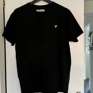 Svart t-shirt från Lyle & Scott - Snygg svart t-shirt från Lyle & Scott med deras ikoniska gula logga på bröstet. Tillverkad i ekologiskt material för en skön och hållbar känsla. Perfekt för en stilren look.
