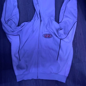 Vit hoodie från Weekday - Säljer en vit hoodie från Weekday med dragkedja och tryck på både fram- och baksidan. Hoodien har svarta detaljer längs ärmarna och en bekväm passform. Perfekt för en avslappnad stil.