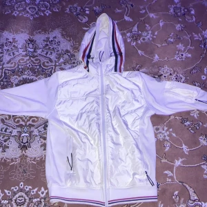 Vit vindjacka från Moncler - Säljer en stilren vit vindjacka från Moncler med dragkedja och huva. Jackan har randiga detaljer i rött och blått på huvan och nederkanten. Perfekt för vår och sommar.