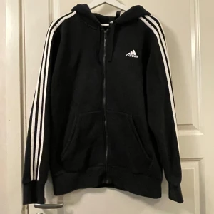 Svart hoodie från Adidas - Svart hoodie från Adidas med dragkedja och de klassiska tre vita ränderna längs ärmarna. Tröjan har en huva och två fickor framtill. Bra skick.