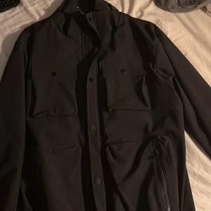 Svart jacka från Zara - Snygg svart jacka från Zara med knappar och dragkedja. Jackan har långa ärmar och flera praktiska fickor framtill. Perfekt för en stilren look.