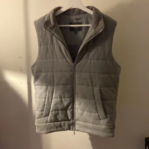 Grå väst  - Säljer en stilren grå väst från Dressmann. Västen har en dragkedja framtill och praktiska fickor. Nypris 900kr. Perfekt för att bara ha över en tröja under kyligare dagar.