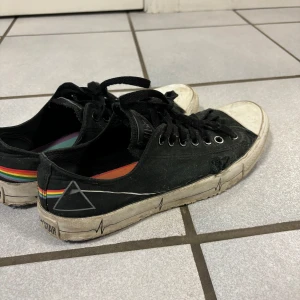 Converse x Pink Floyd  - Unika svarta Converse sneakers med regnbågsfärgad rand och triangeldetalj på sidan. Skorna har snörning och en vit sula med svart mönster. Perfekta för en personlig stil. Hål på högra skon 
