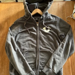 Grå hoodie från True Religion - Säljer en snygg grå hoodie från True Religion med dragkedja och stor logga på ryggen. Tröjan har en justerbar huva och långa ärmar. Perfekt för en avslappnad stil.