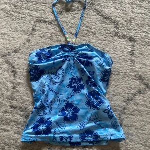 Blå blommig tubtopp med pärldetaljer - Säljer en somrig blå tubtopp med mörkblå blommönster och text. Toppen har en halterneck med färgglada pärlor som detalj. Perfekt för varma dagar!