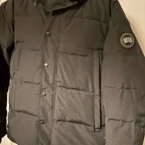 Svart pufferjacka Canada Goose - Säljer en svart pufferjacka från Canada Goose med knappar och dragkedja. Jackan har en broderad logga på ärmen och är perfekt för kalla dagar. Den är långärmad och har en klassisk design som håller dig varm och stilren.