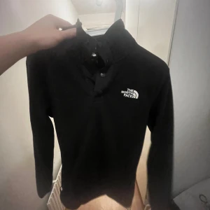 Svart långärmad tröja från The North Face - Säljer en svart långärmad tröja från The North Face med en liten logga på bröstet. Perfekt för kyligare dagar och passar till det mesta. Tröjan har en klassisk design med knappar vid halsen.
